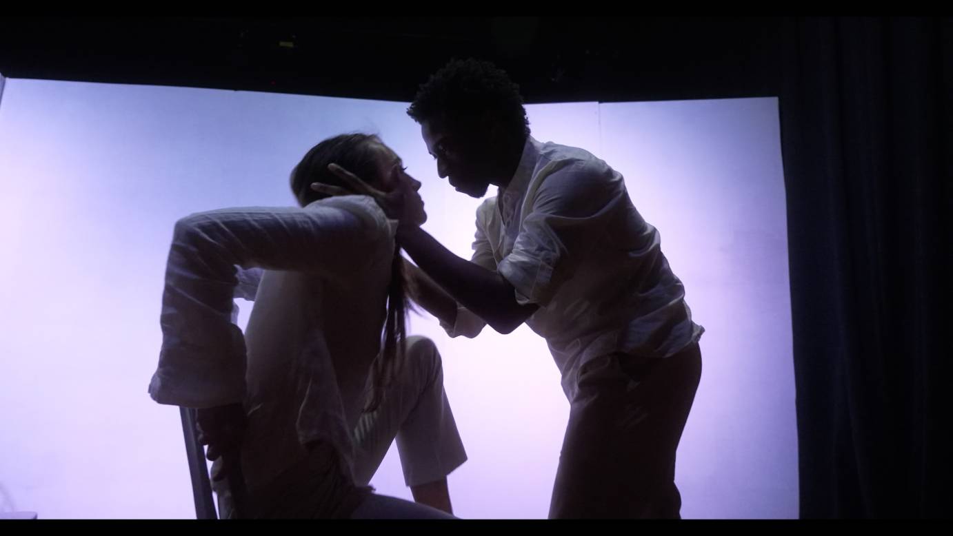 under purple pink light a man grabs a seated womans face and gazes at her intently